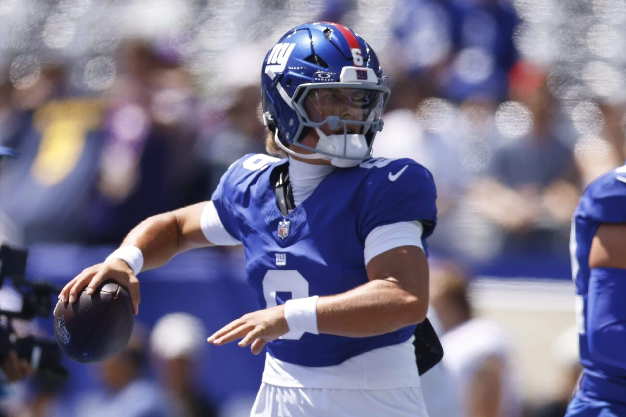 Jaxson Dart is shown from the waist up, preparing to throw with the football in his right arm and his left arm in front of him. He's wearing a blue Giants jersey and helmet.