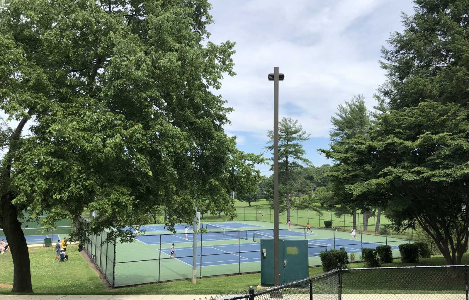 Outdoor park scene with blue tennis courts, players, and a chain-link fence framed by large green trees under a partly cloudy sky