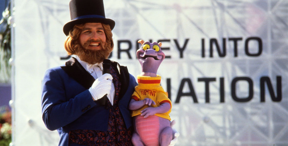 Dreamfinder, a man in a tuxedo and top hat, holds Figment, a small purple cartoon-like dragon wearing a yellow sweater, standing in front of a sign that partially reads