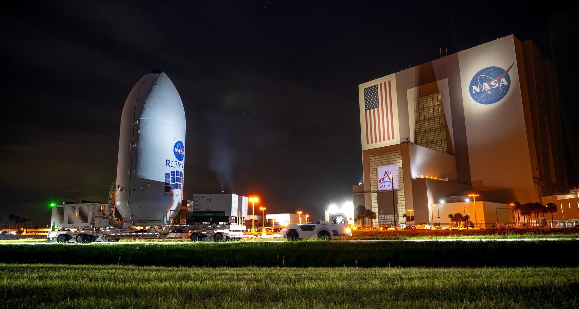 A large rocket payload fairing labeled “NASA Roman Space Telescope” is transported on a flatbed trailer at night near NASA’s Vehicle Assembly Building at Kennedy Space Center. The illuminated VAB, displaying a large American flag and NASA logo, stands in the background under a dark sky.