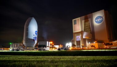 A large rocket payload fairing labeled “NASA Roman Space Telescope” is transported on a flatbed trailer at night near NASA’s Vehicle Assembly Building at Kennedy Space Center. The illuminated VAB, displaying a large American flag and NASA logo, stands in the background under a dark sky.