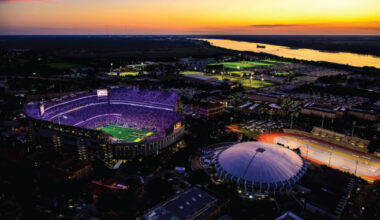 Aerial view of LSU Tiger Stadium at dusk, glowing purple and surrounded by a circular domed arena, with a river in the distance.