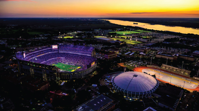 LSUCampus2_LSUAthletics Aerial view of LSU Tiger Stadium at dusk, glowing purple and surrounded by a circular domed arena, with a river in the distance.