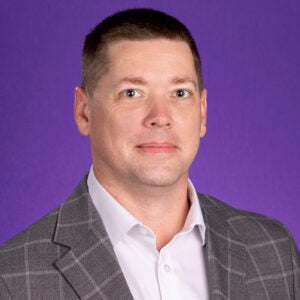 Head-and-shoulders portrait of a person with short dark hair wearing a gray plaid blazer over a light-colored collared shirt. They face the camera with a slight smile against a solid purple studio background.