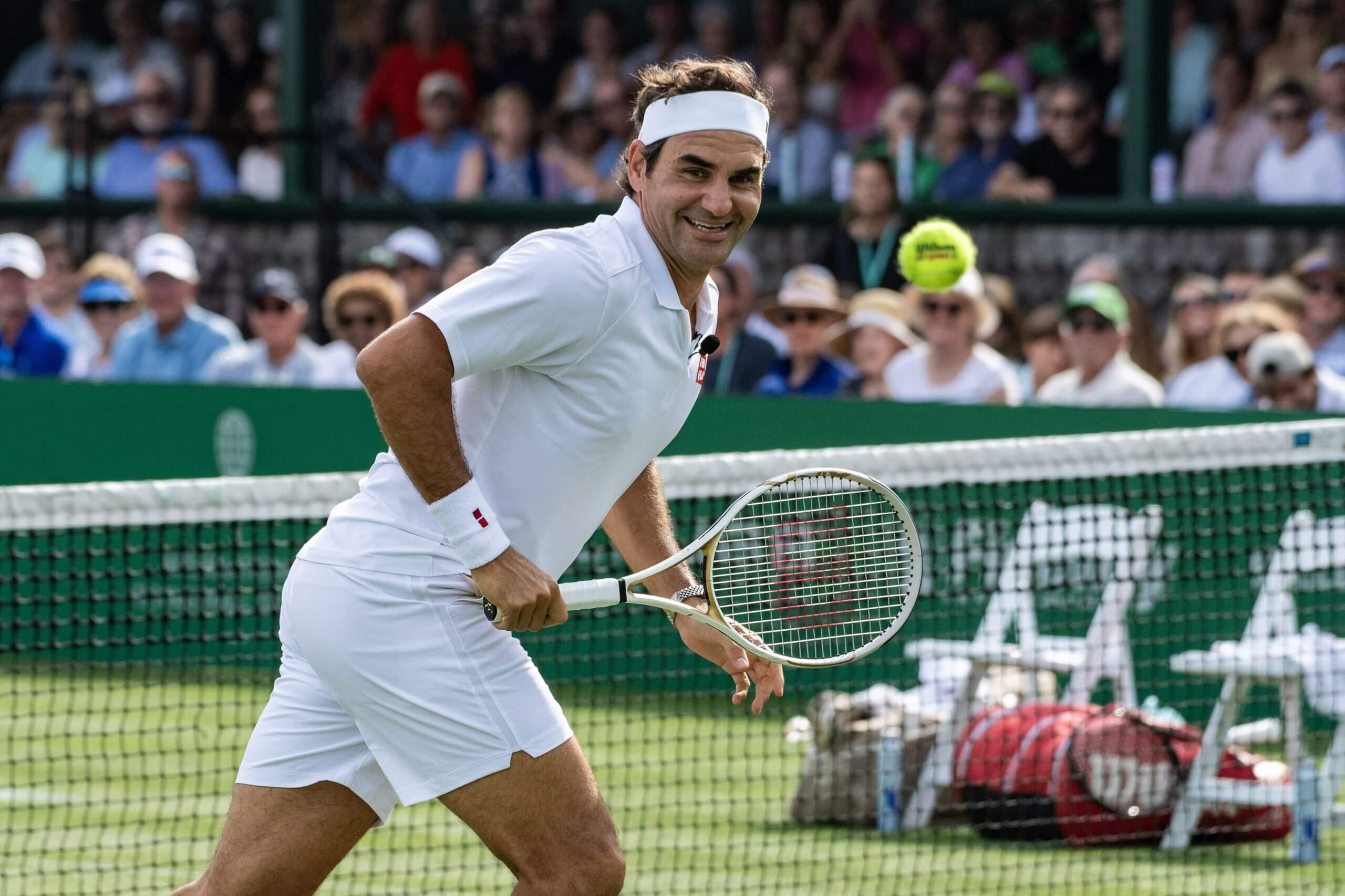 Roger Federer looks on at a tennis ball floating behind him on a grass court with a crowd in the background.