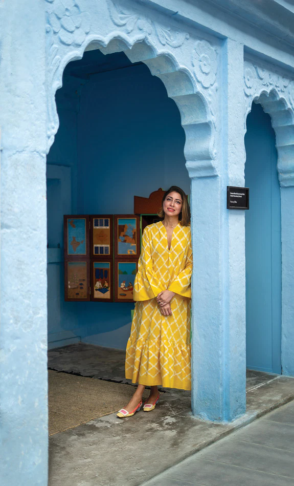 Person in a yellow dress standing in a blue architectural setting with a framed artwork on the wall in the background.