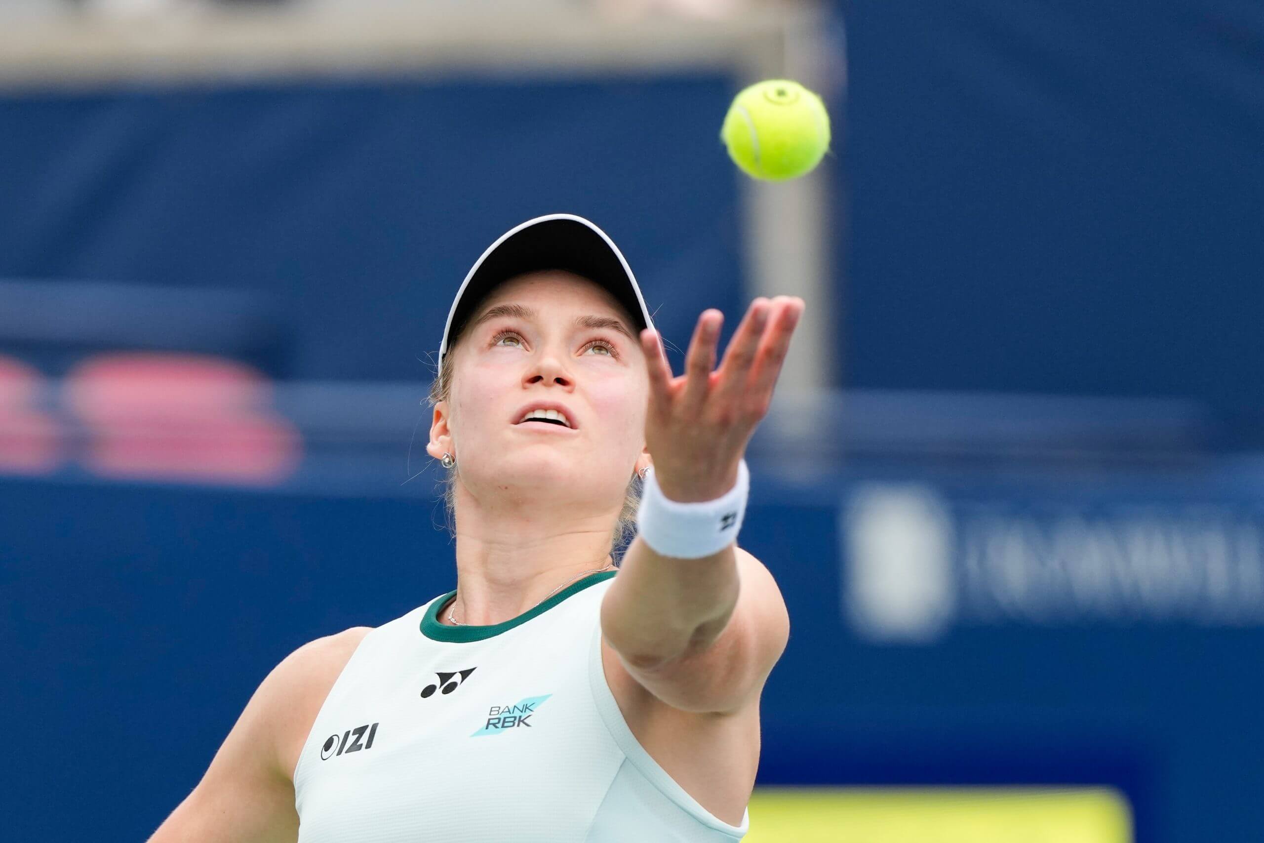 Elena Rybakina tosses a tennis ball into the air with her left hand.