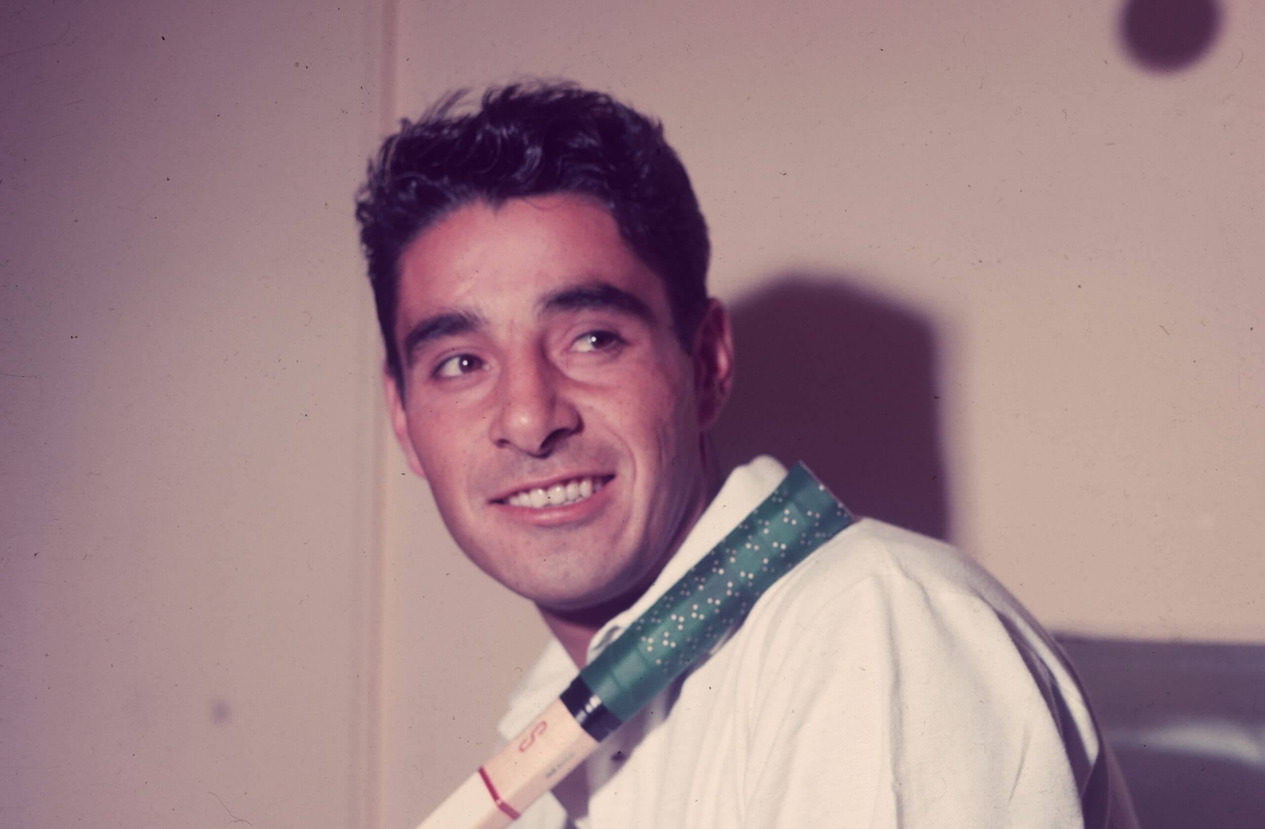 Pancho González sits looking to his left with a tennis racket handle resting across his left shoulder.