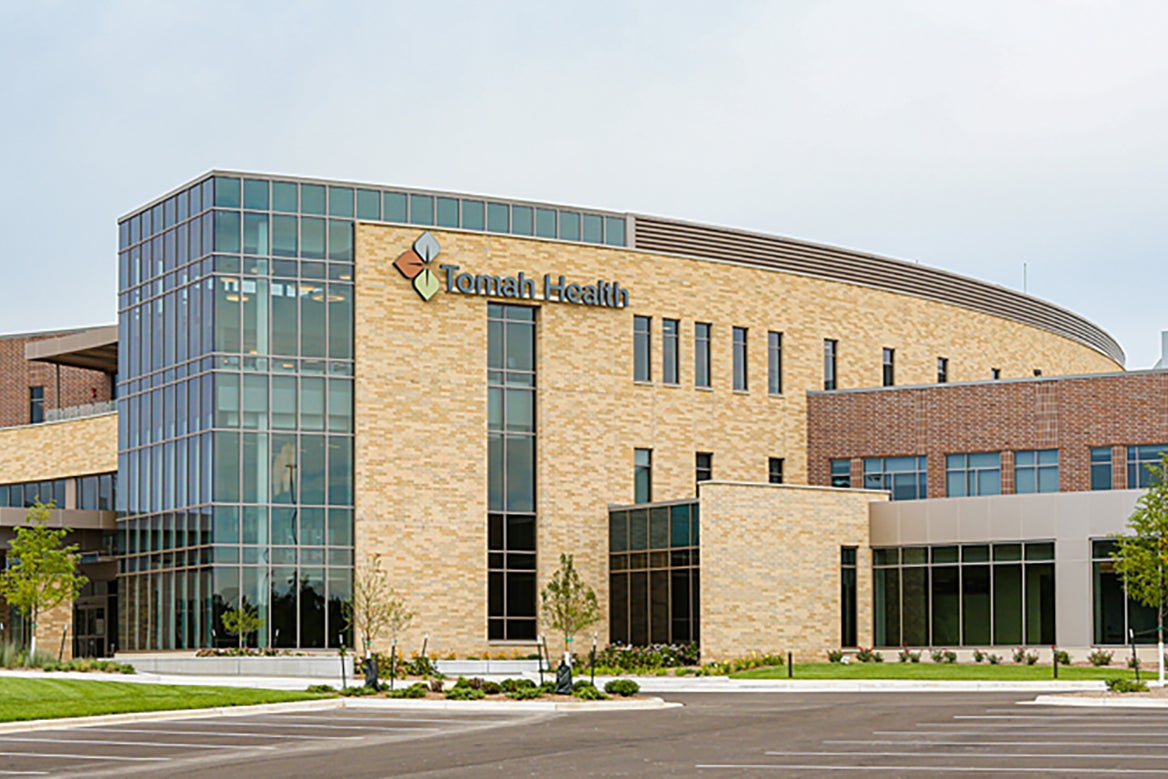 Modern healthcare building with large glass windows, a tan brick facade, and Tomah Health signage on the exterior; trees and an empty parking lot in front.