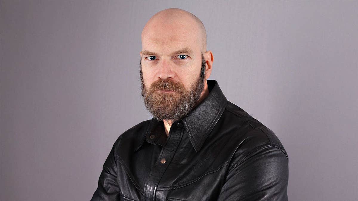 Tyler Mane posing in a leather jacket against a light purple backdrop.