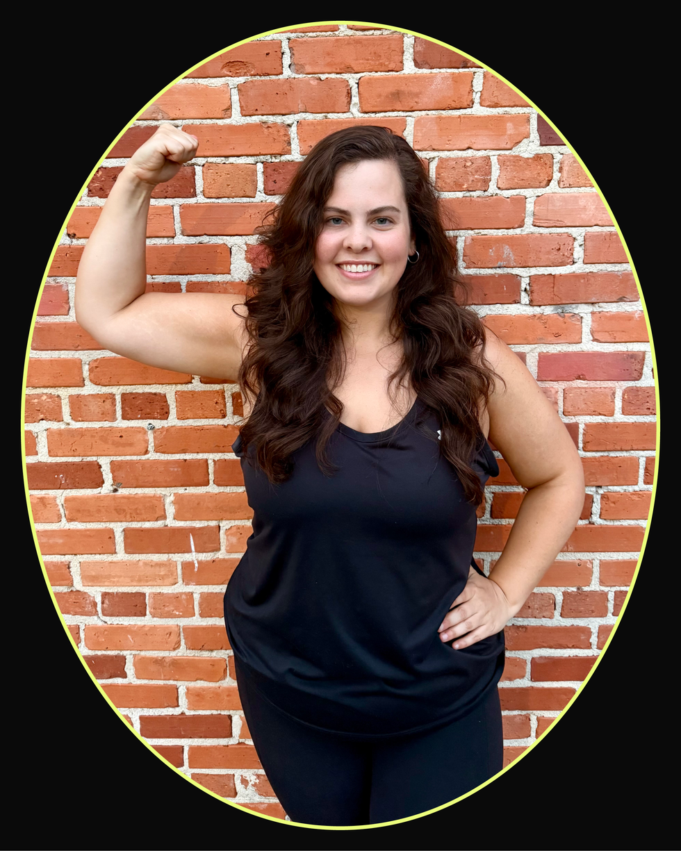 Smiling woman in a black tank top flexing her arm against a brick wall background.