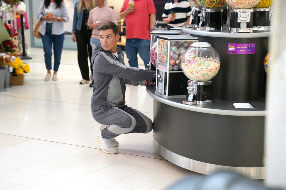 hollyoaks ste hay person in tracksuit kneeling near candy vending machine in a mall.