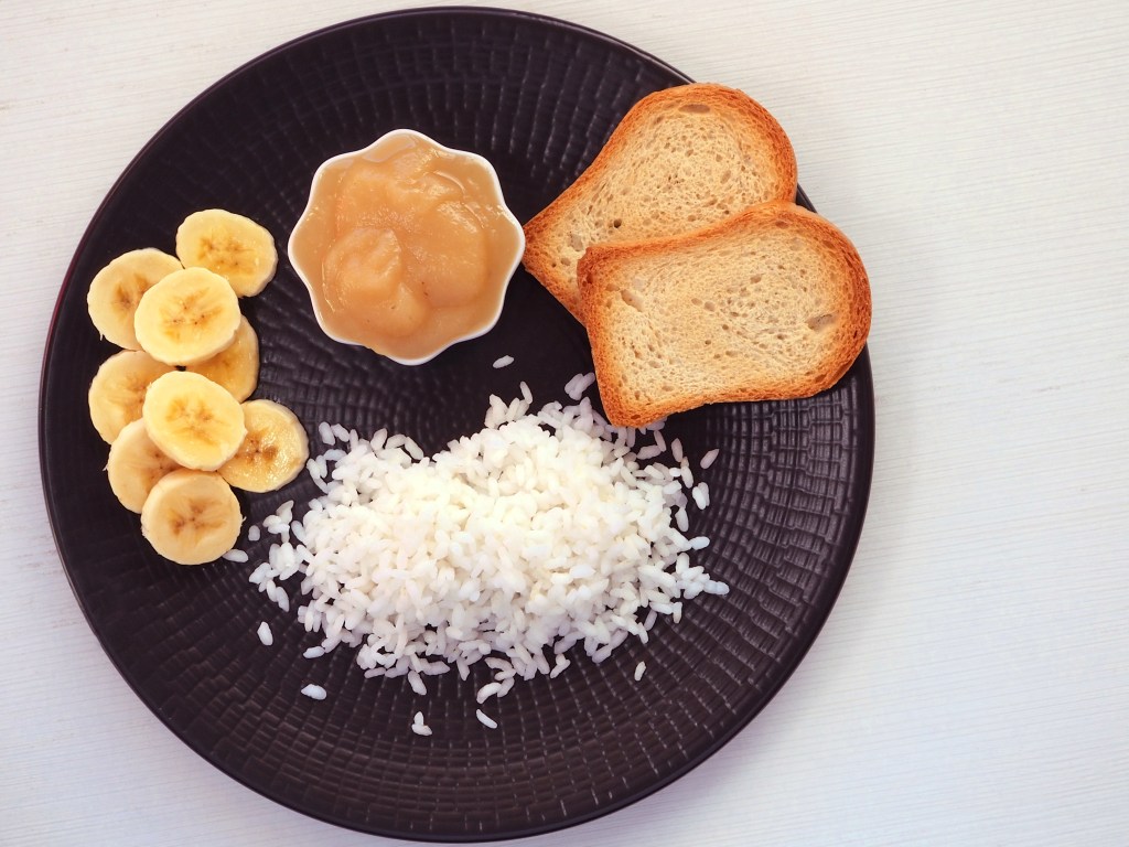 Banana slices, boiled rice, applesauce, and toast on a black plate.