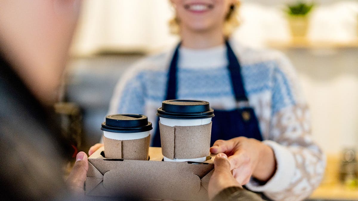A barista smiles while she hands two coffees in to-go cups to a customer.