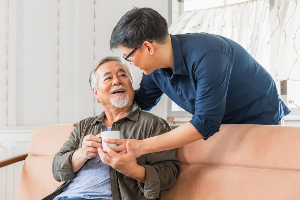 A middle-aged man giving a senior man a cup of coffee on the couch.