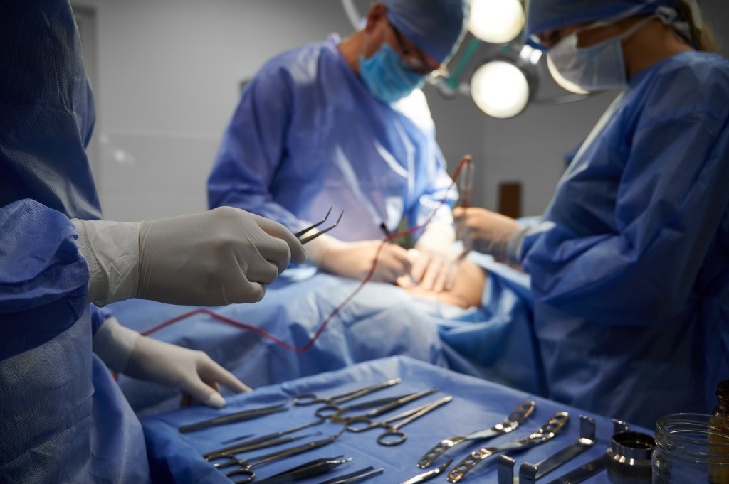 A close-up of a gloved hand holding tweezers over a tray of surgical instruments while a surgical team performs plastic surgery in the background.