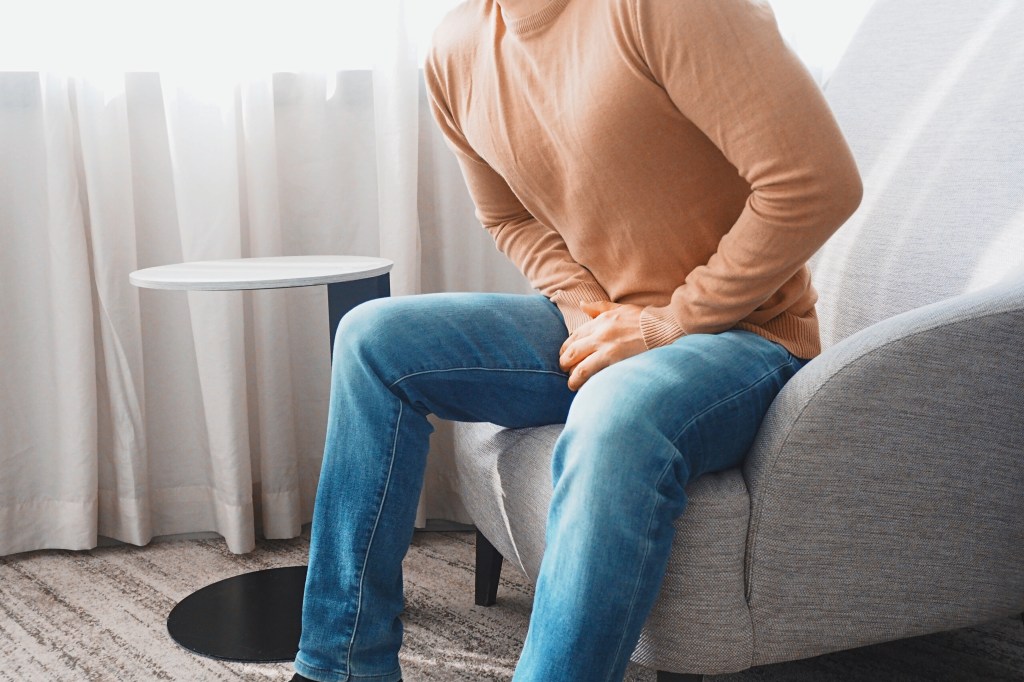 A man sits on a chair, holding his crotch due to pain from a genitourinary problem.