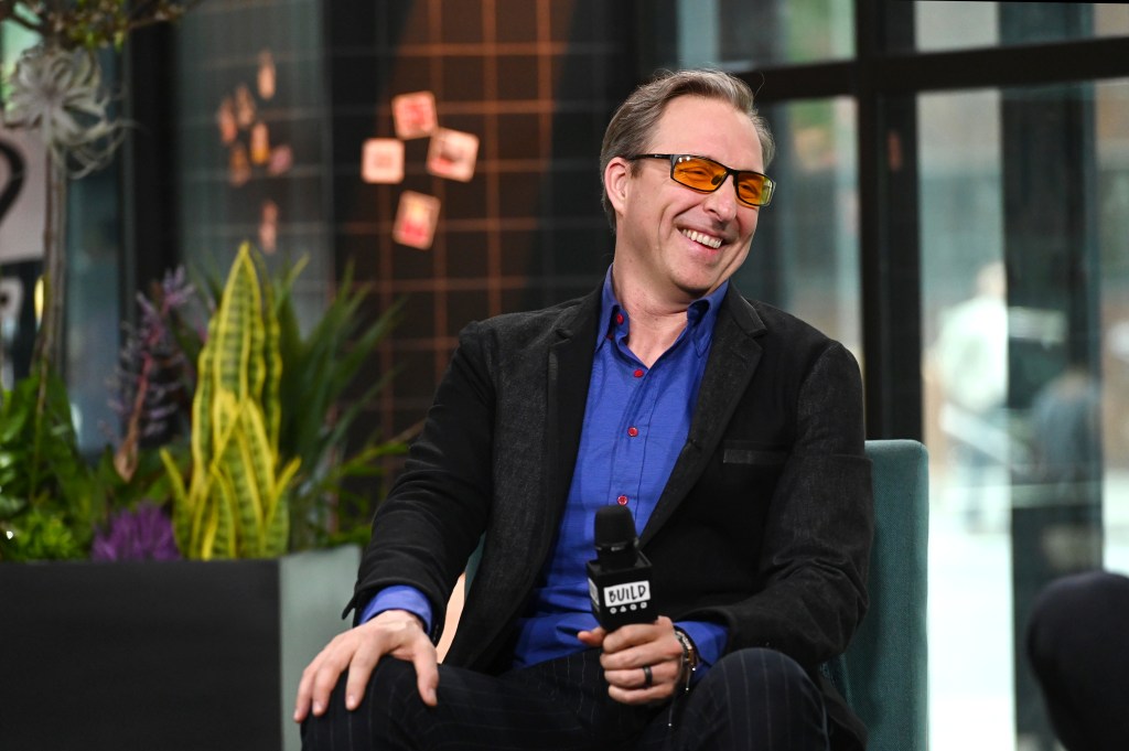Dave Asprey in a black jacket and blue shirt, wearing orange glasses and smiling, holding a "BUILD" microphone.