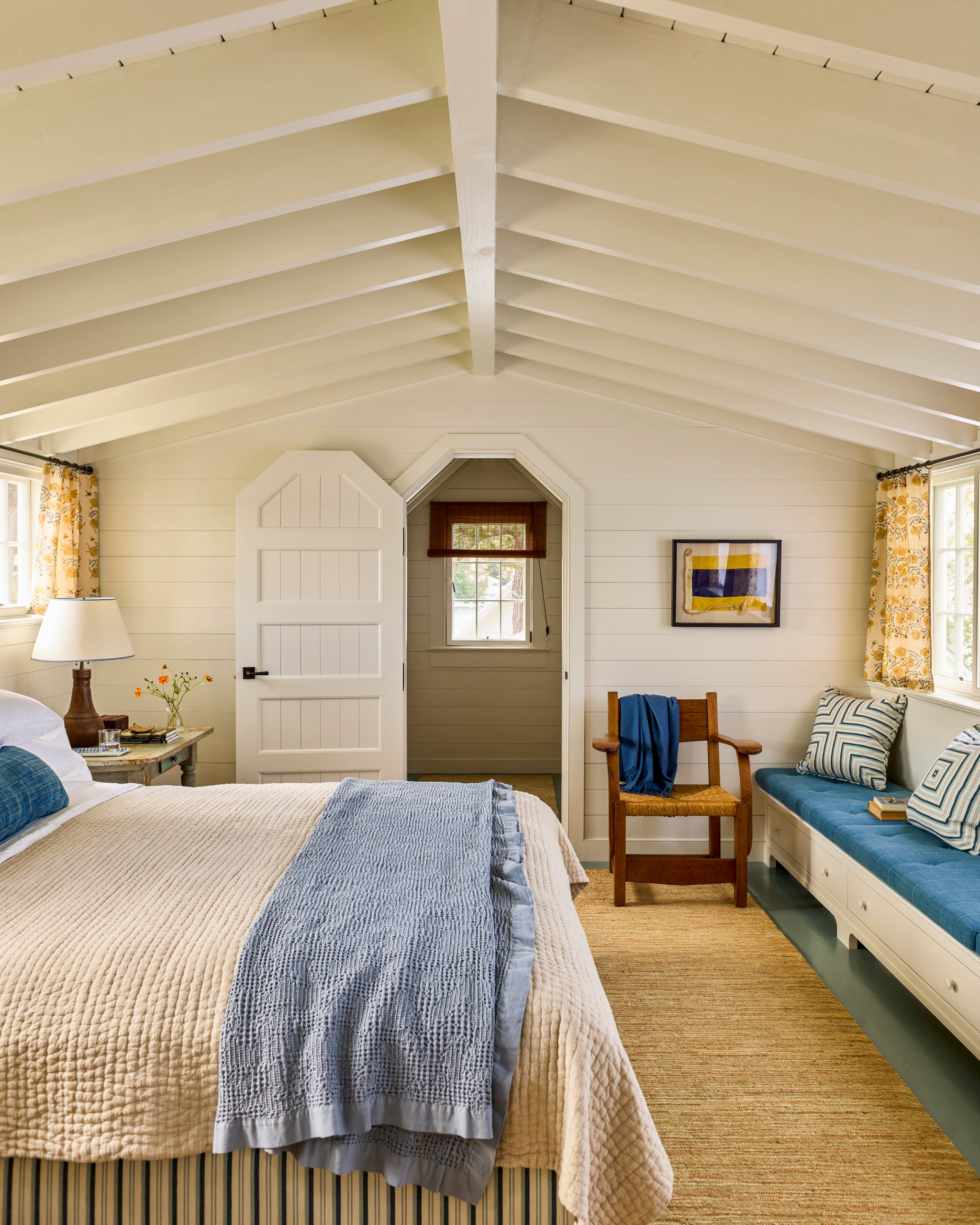 New Hampshire Lake Cottage by Schafer & Company Bedroom with a bed, cozy seating area, wooden chair, and bright windows, featuring vaulted ceilings and warm decor.