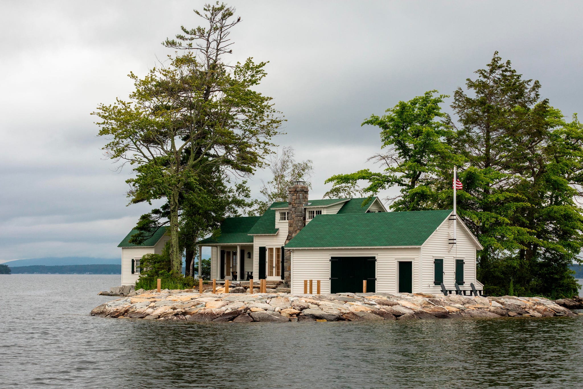 Island lake house with a green roof situated on a rocky shoreline, surrounded by trees and overlooking the water.