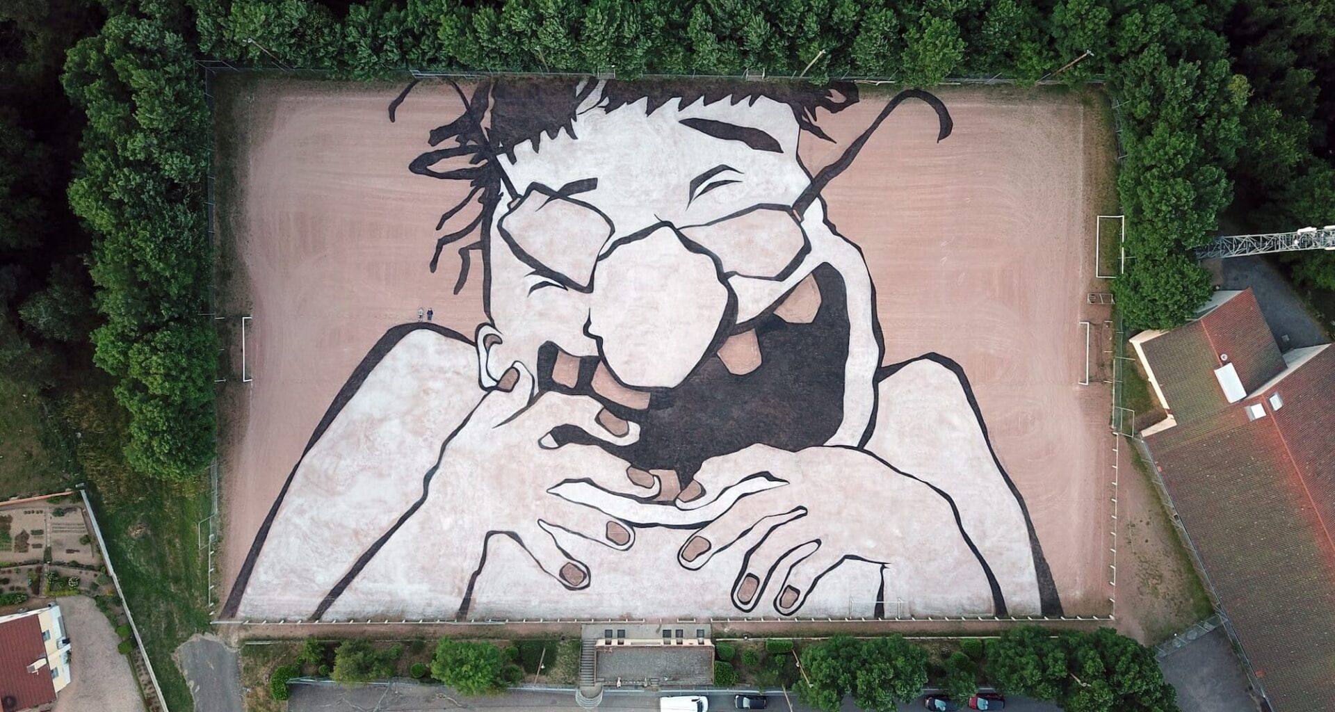 an aerial view of a large mural by Ella & Pitr on a playing field featuring a cartoonish laughing figure