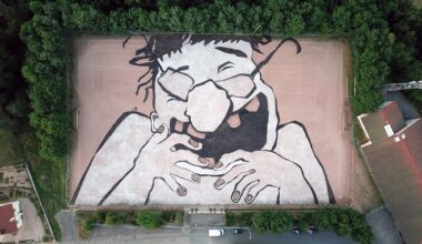 an aerial view of a large mural by Ella & Pitr on a playing field featuring a cartoonish laughing figure