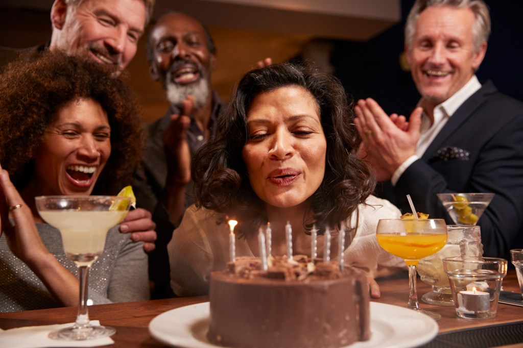 Woman blowing out candles on a chocolate birthday cake with friends clapping and celebrating in a bar.