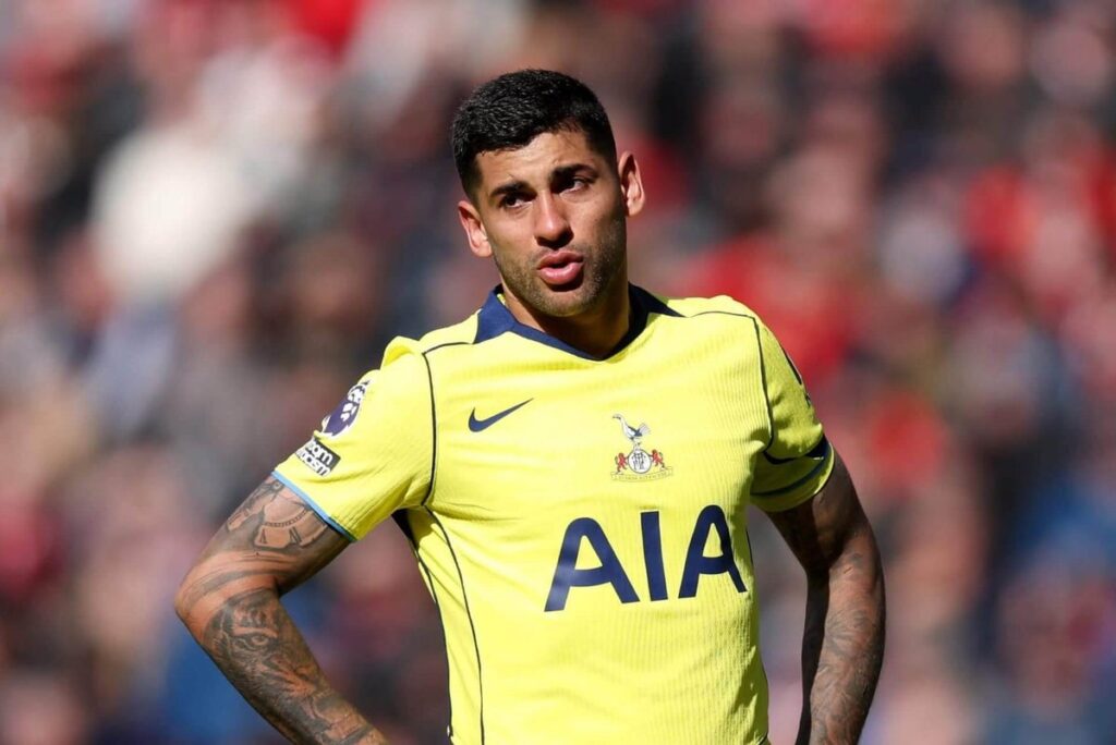 Cristian Romero, wearing a yellow Tottenham shirt, stands with his hands on his hips.