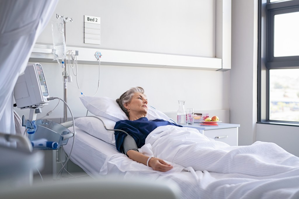 A lonely elderly woman lies in a hospital bed, looking away thoughtfully.