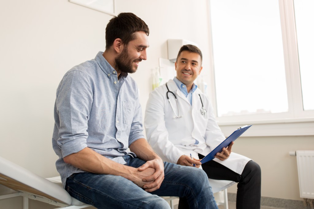 A smiling doctor holding a clipboard talks with a young male patient.
