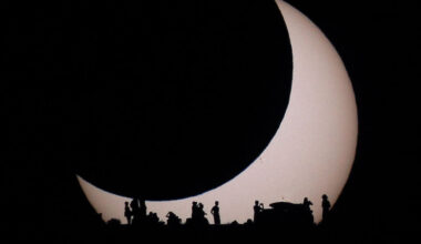 A light disk is visible on the lower left but the
rest of the disk is eclipsed by a darker disk of the 
same apparent size. At the bottom of the image are the
silhouettes of several people. 
Please see the explanation for more detailed information.