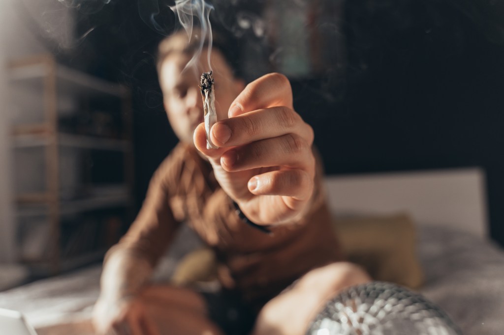 A man holding out a lit, hand-rolled cannabis cigarette.