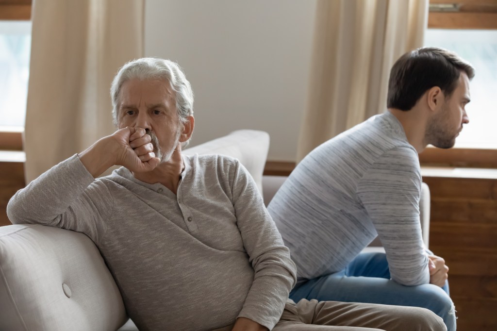 Unhappy father and adult son sitting back-to-back on a couch after a quarrel.