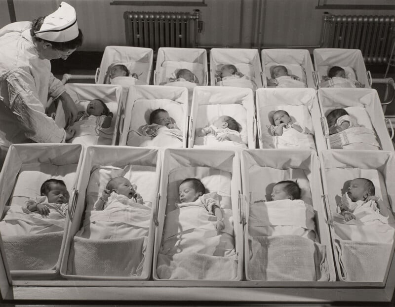 A nurse in a white uniform and cap tends to a row of newborns sleeping in individual hospital bassinets.