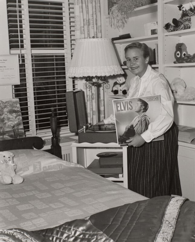 A young woman smiles while holding an Elvis Presley vinyl record in a bedroom with a record player, a bed, and shelves filled with stuffed animals.