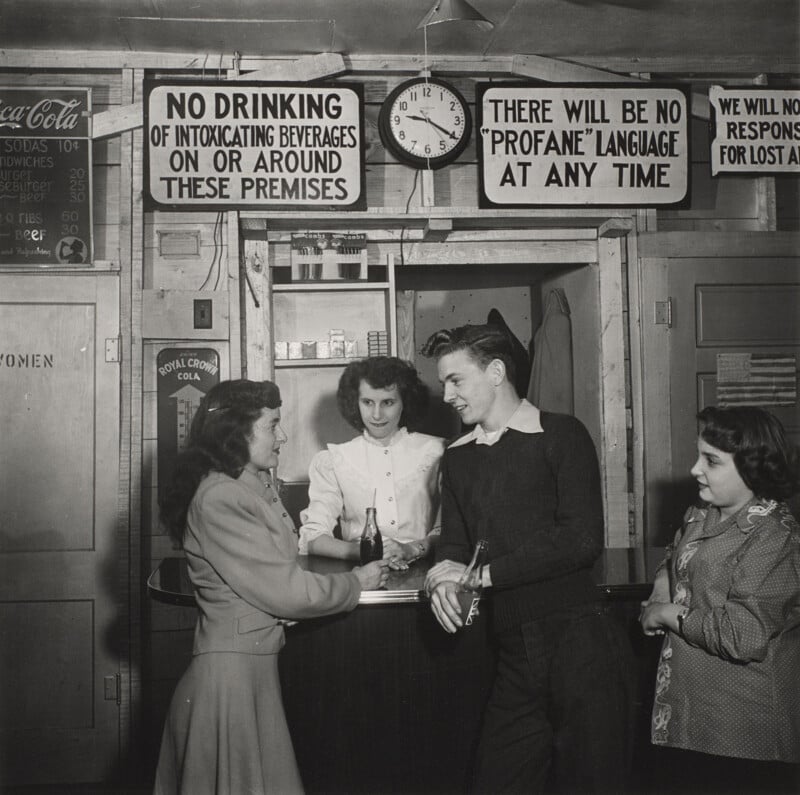 Four young people stand around a counter in a room with signs prohibiting alcohol and profanity, while a menu lists food prices.