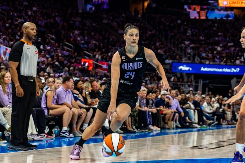 A basketball player in a Valkyries jersey dribbles the ball on court during a game, with a referee and spectators visible in the background.