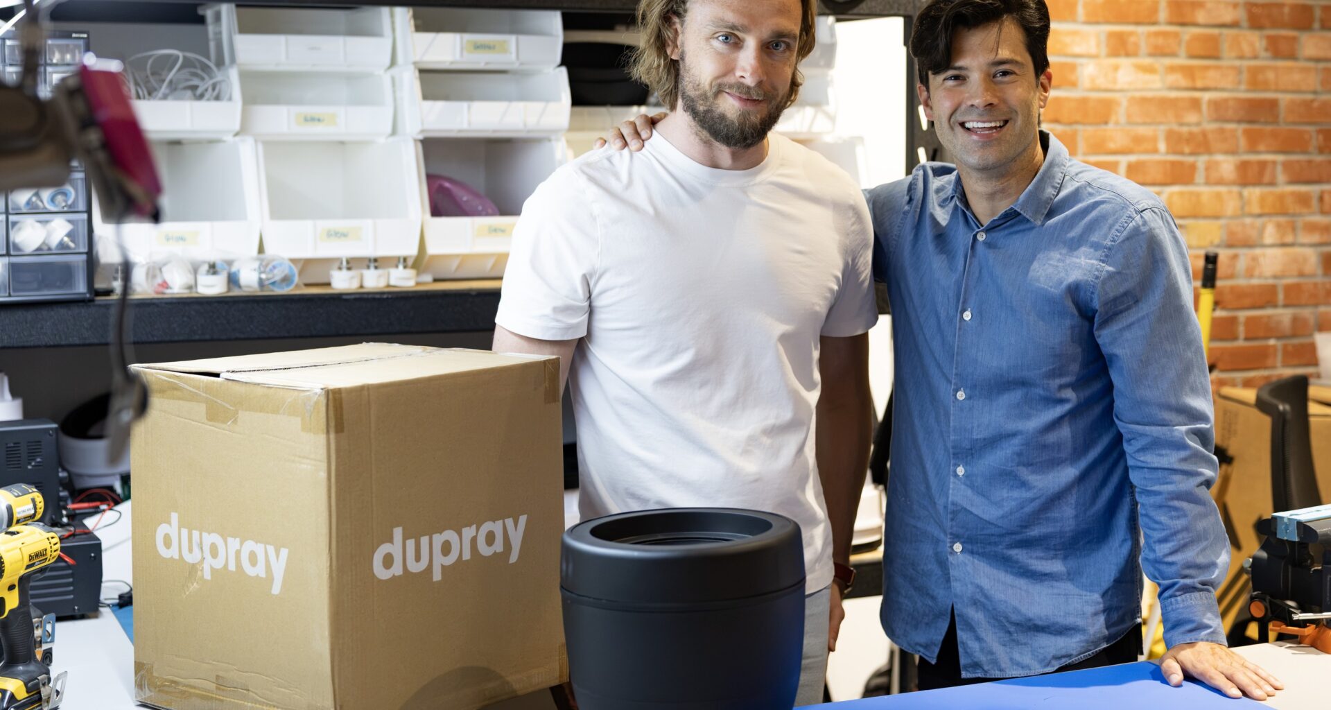 Two guys standing beside a box with their company name on it and a small, portable humidifier on a table in front of them.