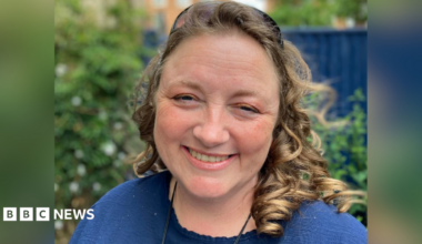 A woman with curled hair smiling into the camera. She is wearing a blue top and is stood in a garden, with the background slightly blurred.