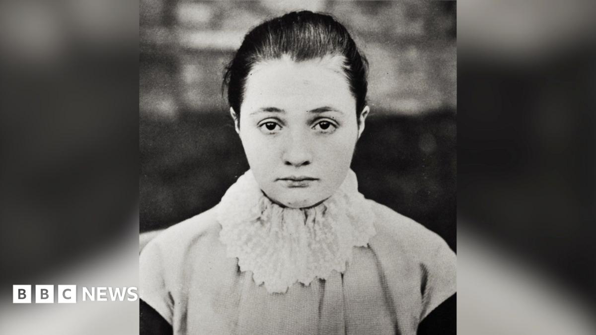 A black and white photograph of a young woman with hair tied back, looking straight at the viewer. She is wearing a white, frilly ruffle over a light-coloured top.