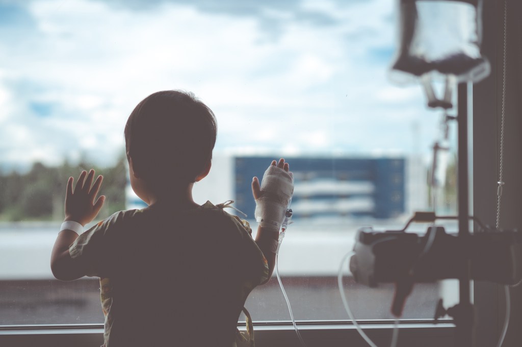 Toddler boy with IV tube looking out hospital window.