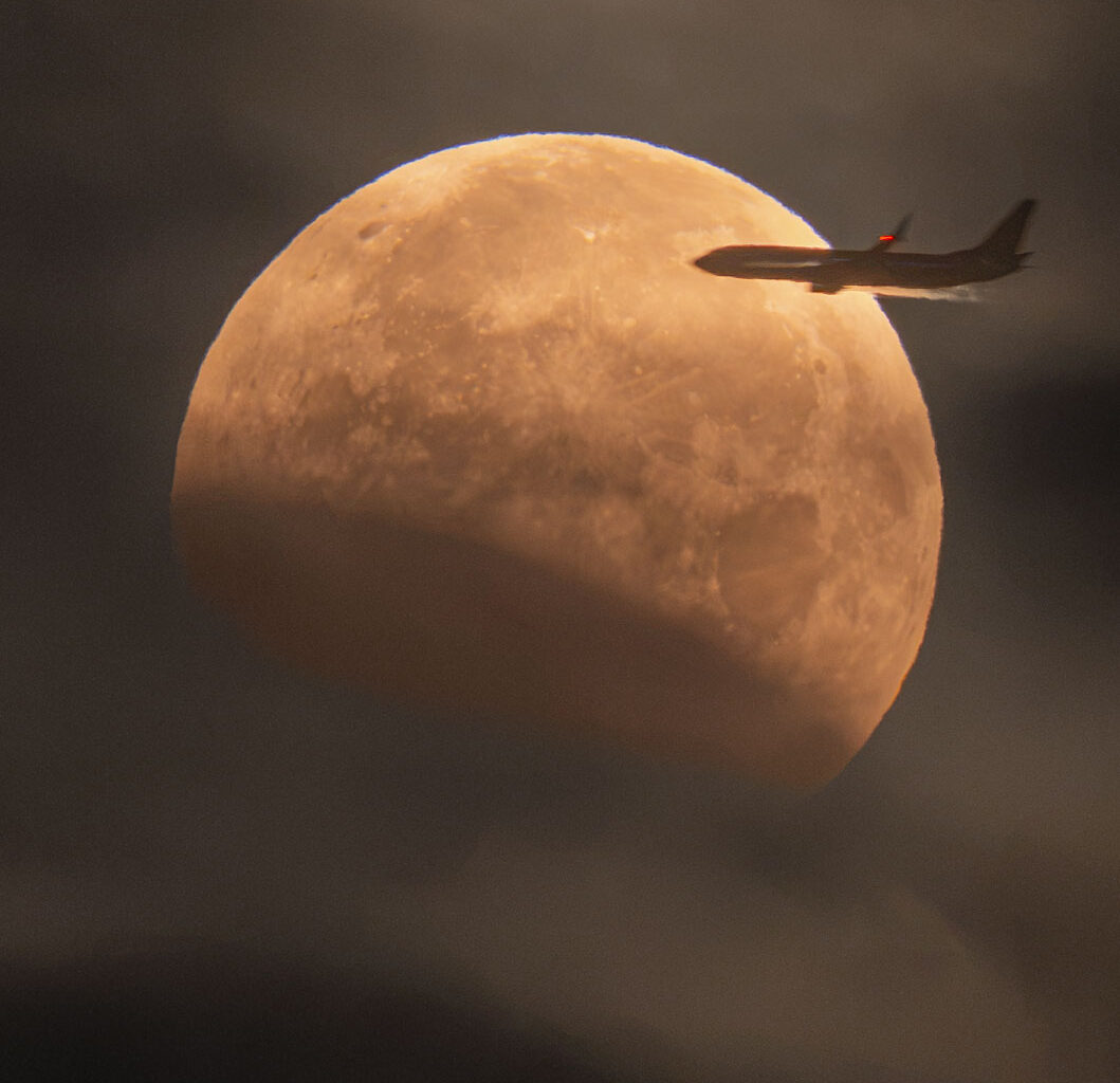 The Moon is seen in front of clouds but with part
of its lower left being unusually dark. On the upper 
right, a foreground airplane appears flying into the disk of
the Moon.  
Please see the explanation for more detailed information.