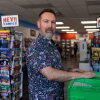 Marc Bowker, owner of Alter Ego Comics in Lima, Ohio, thumbs through a green bin of comic books and either side of him are two shelves with signs that read "Hey!! Comics." He has short brown hair and a graying beard. He is wearing a patterned button up shirt that has "Avengers" written on it along with Marvel characters such as Iron Man and Captain America.