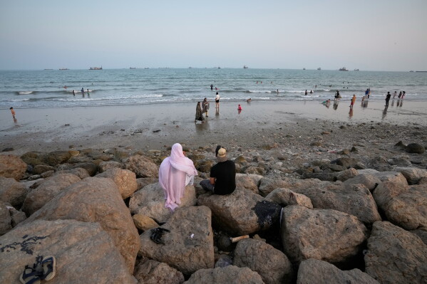Two people sit on rocks overlooking a beach as others swim and commercial vessels lie anchored in the Strait of Hormuz off Bandar Abbas, Iran, Friday, Sept. 4, 2026. (AP Photo/Vahid Salemi)