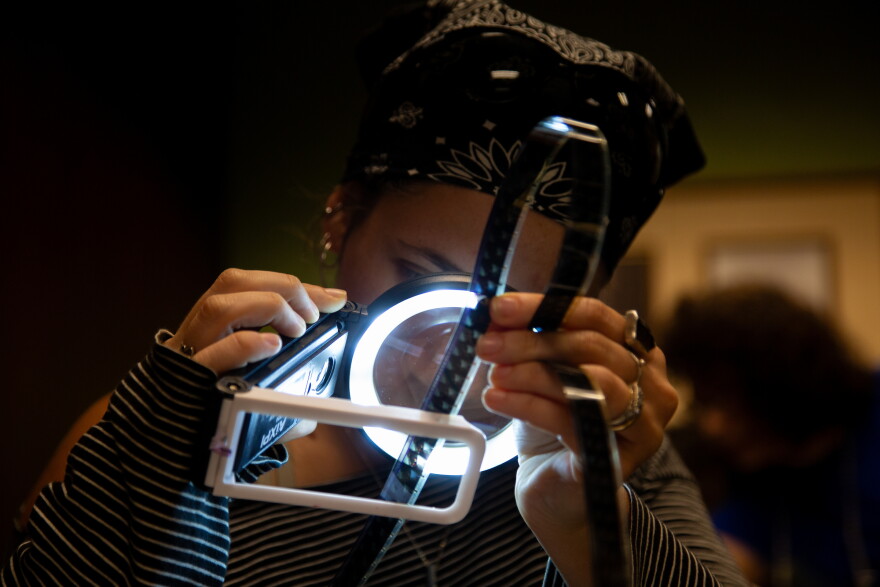 Tess Tarses, 24, picks up a magnifying glass to closely observe developed film at the Film Strip Club meeting at the Howson Branch Library on Monday, August 10, 2026. This is the first Film Strip Club meeting Tarses attended. Ysa Mendoza/KUT