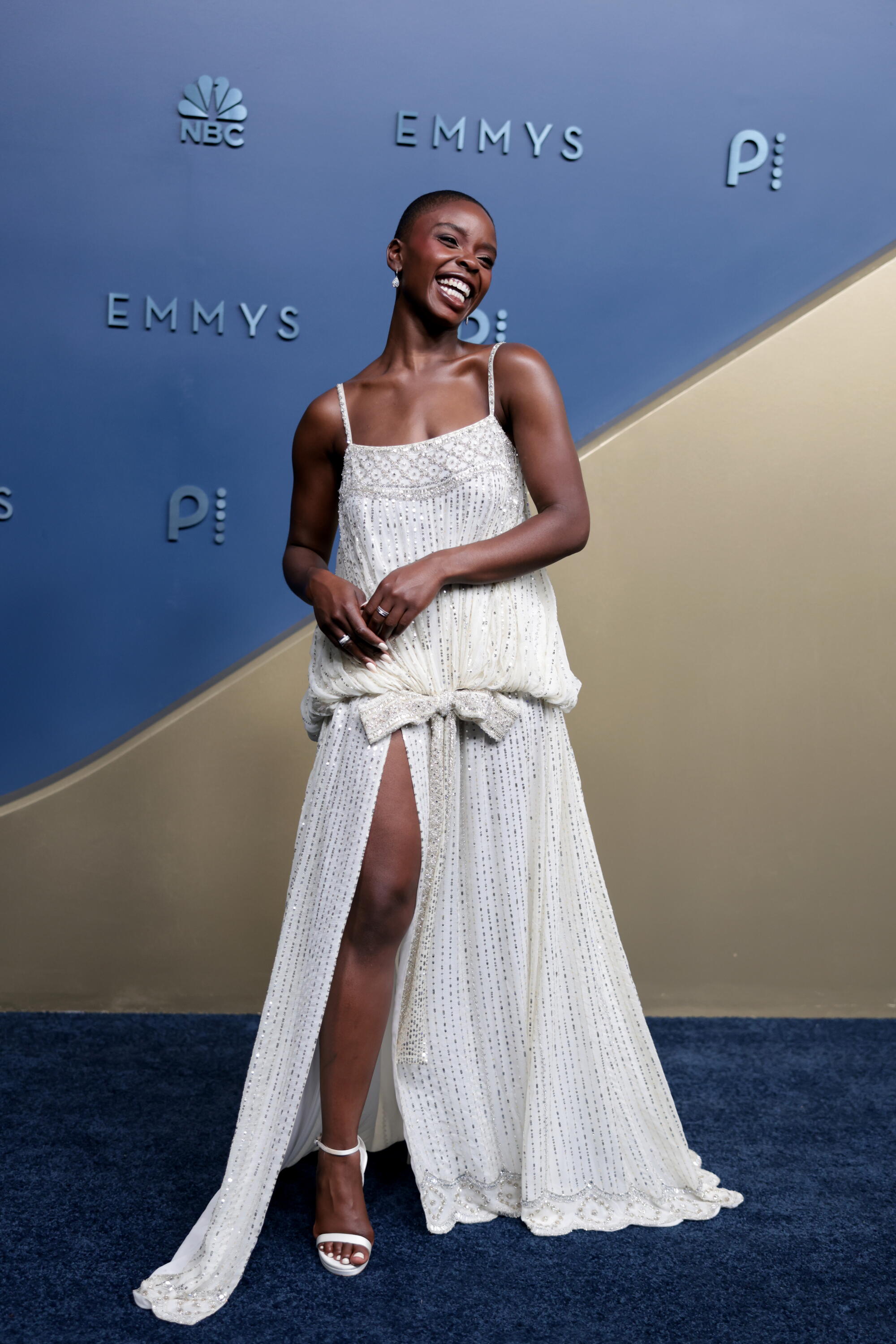 Joy Sunday, wearing a white dress with a high slit, poses on the Emmys blue carpet. 