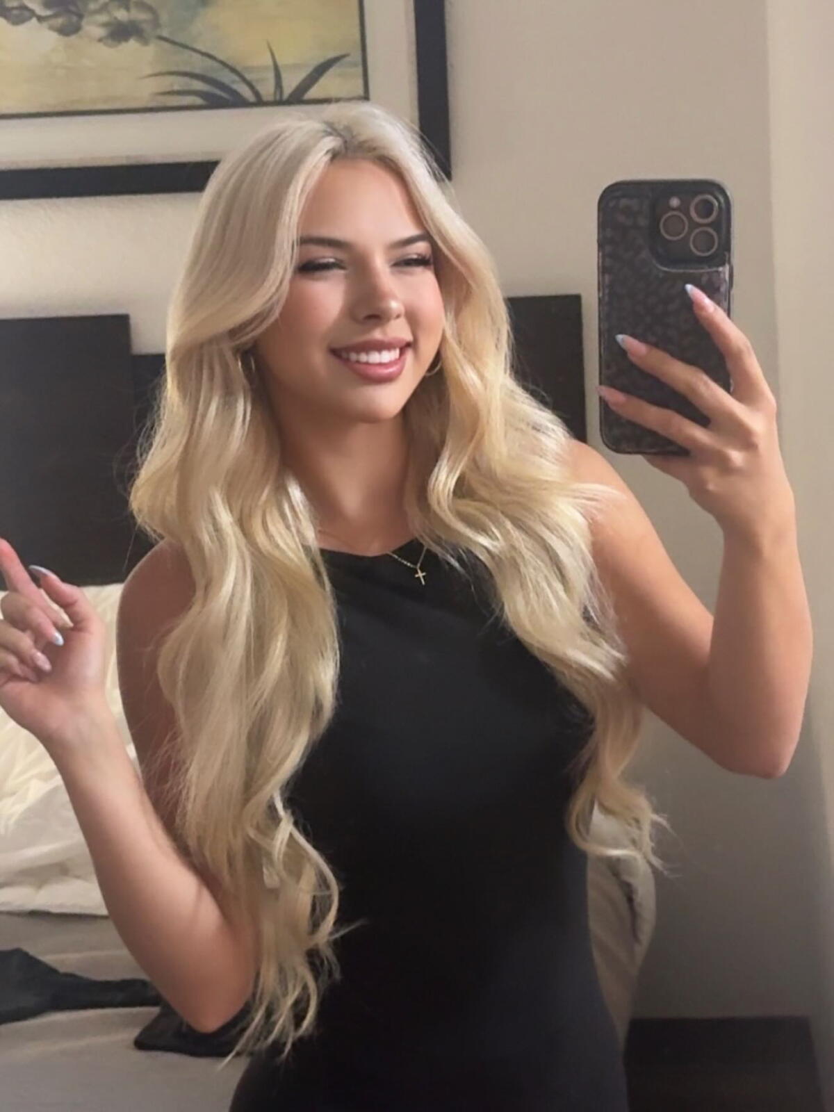 A young woman wearing a sleeveless black dress holds up a cellphone pointed toward her.