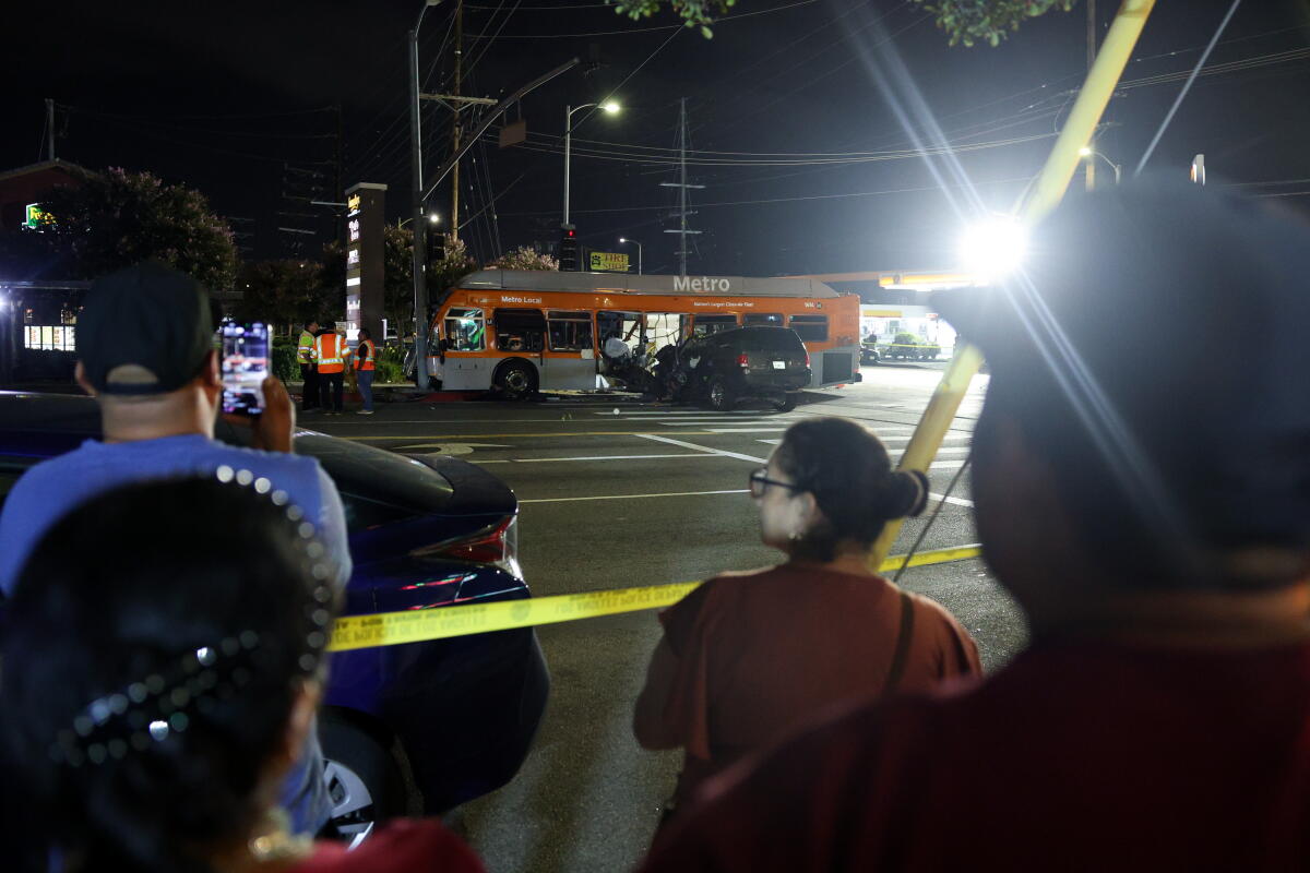 People stand behind yellow tape viewing the scene of a bus crash.