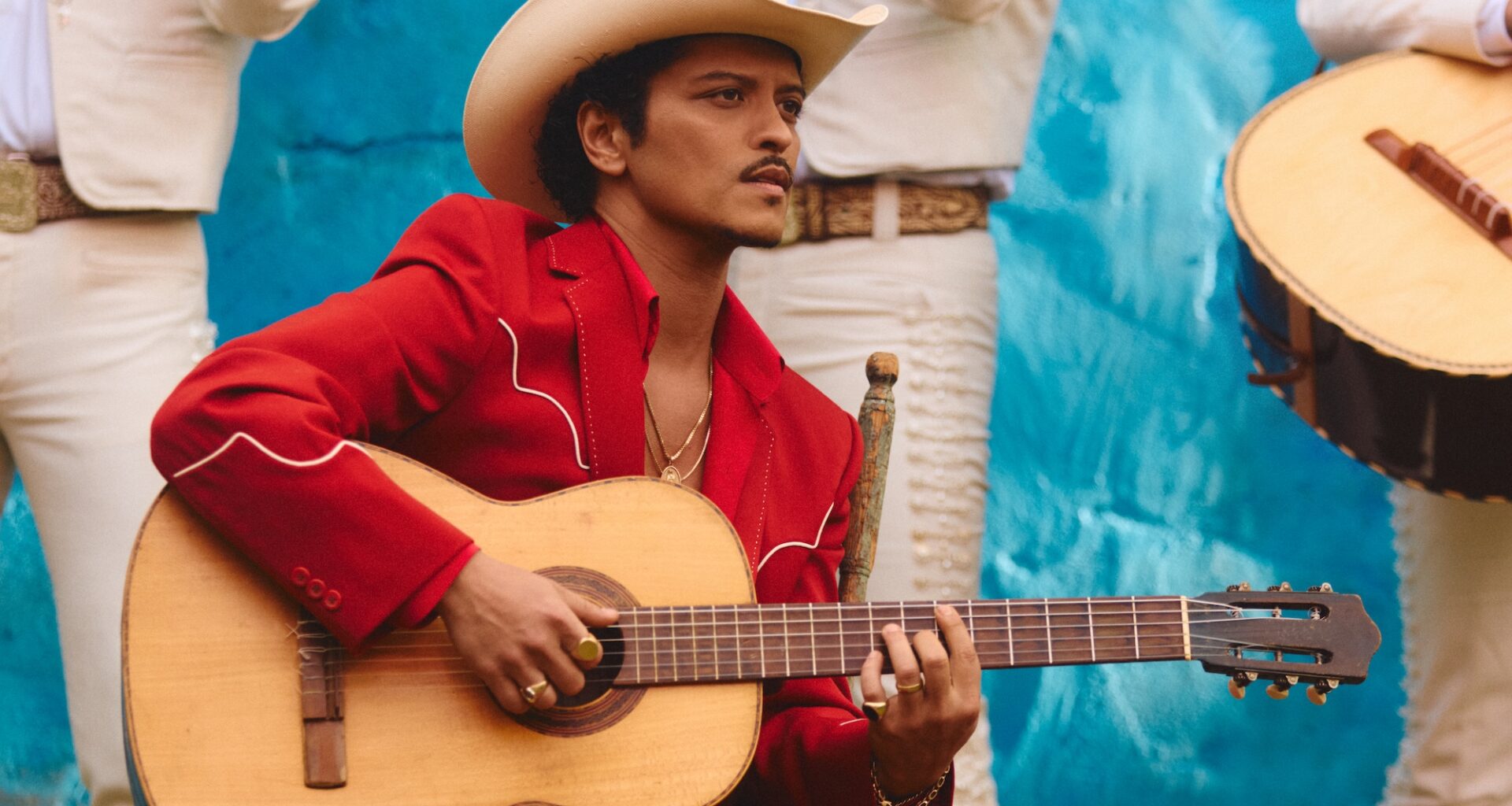 Photo of Bruno Mars wearing a red suit, a white hat, and holding a guitar.