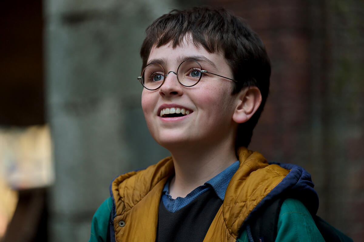 Dominic McLaughlin smiles as HBO's Harry Potter, wearing round glasses and well-worn clothes.